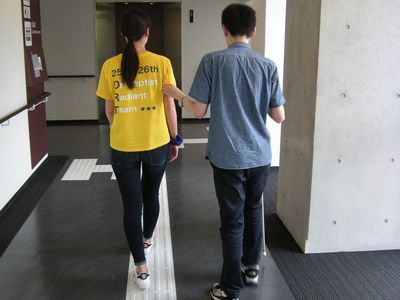 ボランティアの右腕に掴ってもらい建物内を歩いている写真。二人とも後姿が写っている。